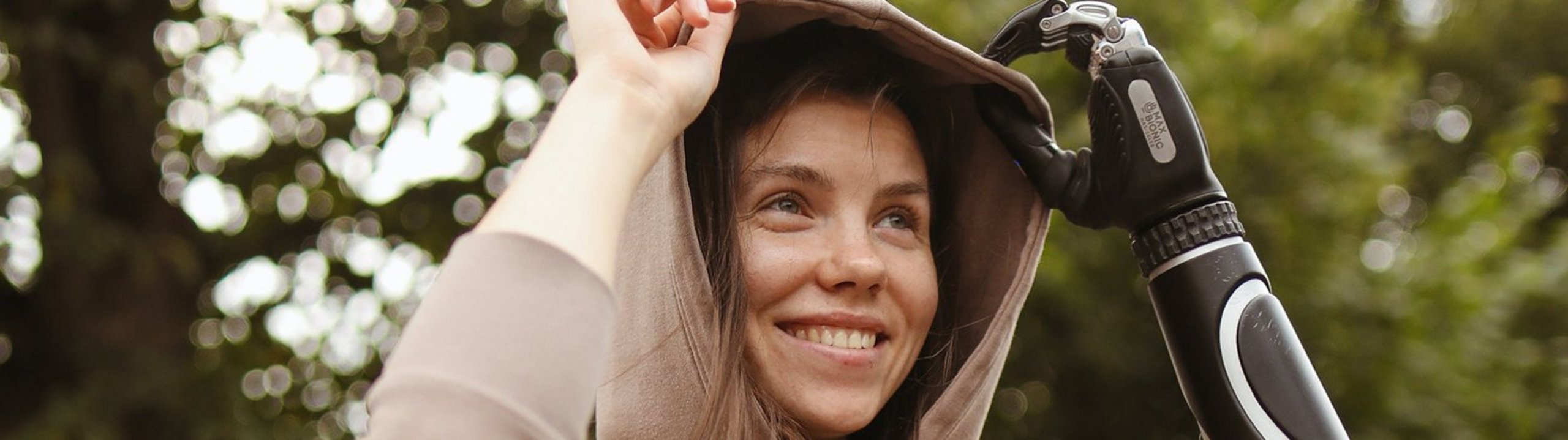 Junge Frau mit Armprothese lächelt und richtet ihre Kapuze im Grünen.