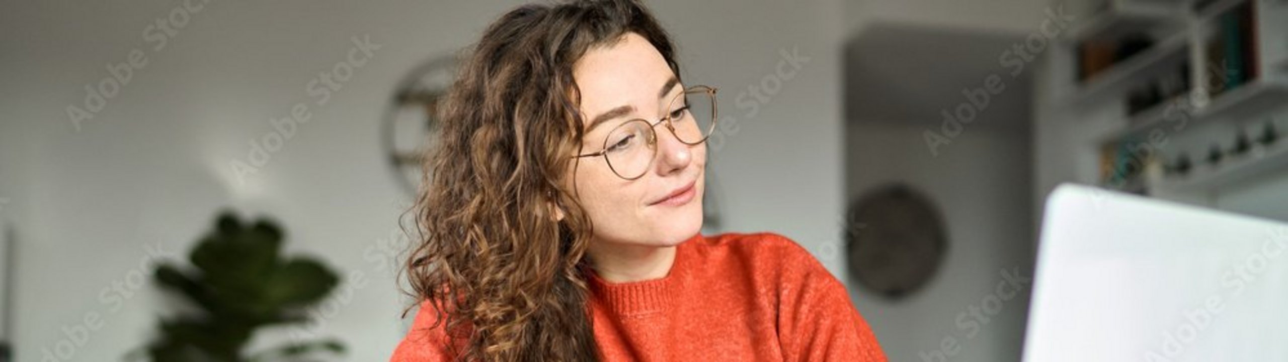 Eine Frau sitzt am Tisch, schreibt in ein Notizbuch und blickt dabei auf ihren Laptop.