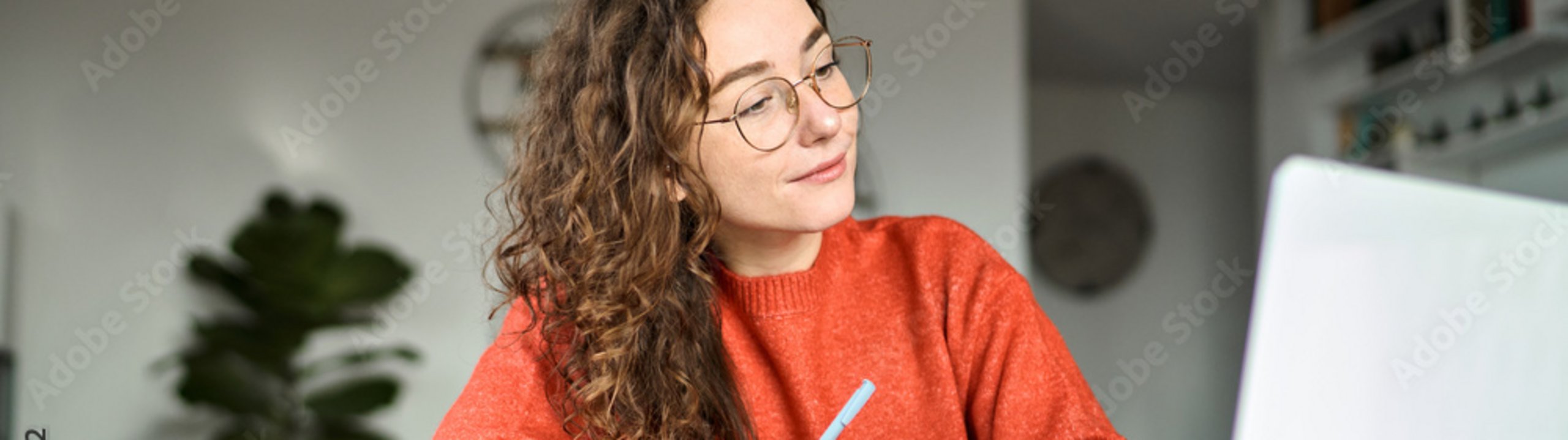 Eine Frau sitzt am Tisch, schreibt in ein Notizbuch und blickt dabei auf ihren Laptop.