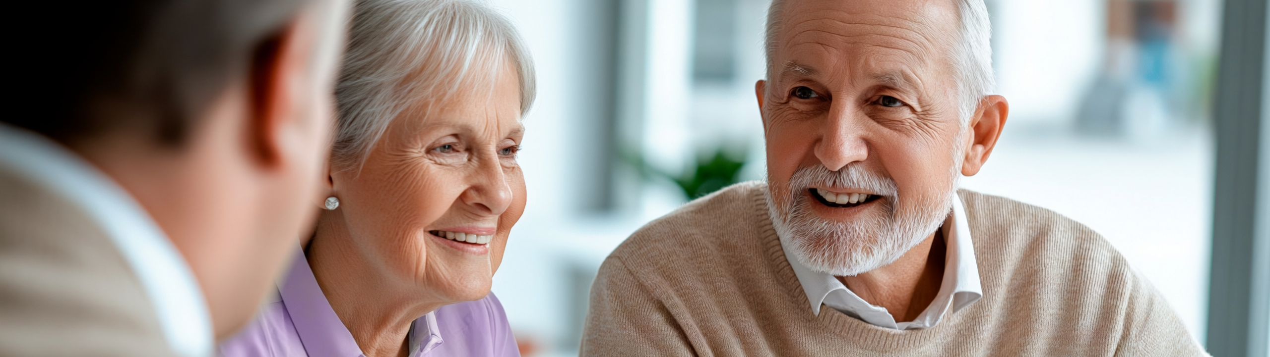 Ein älteres Paar sitzt lächelnd im Gespräch mit einer beratenden Person an einem Tisch.