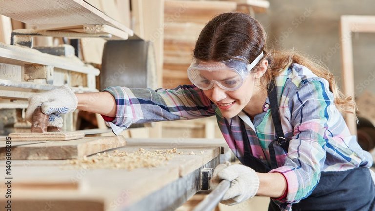 Frau mit Schutzbrille bearbeitet Holz in einer Werkstatt.