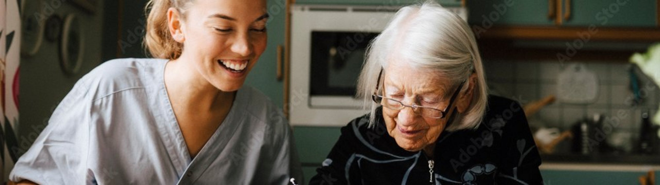 Pflegerin hilft älterer Frau beim Kreuzworträtzel am Küchentisch.