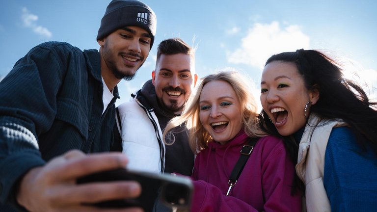 Vier junge Erwachsene lachen und machen gemeinsam ein Selfie im Freien.