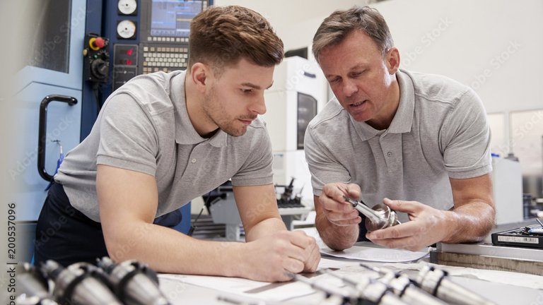 Zwei Personen begutachten gemeinsam ein Metallbauteil und besprechen dessen Bearbeitung an einem Arbeitsplatz in einer Werkstatt.