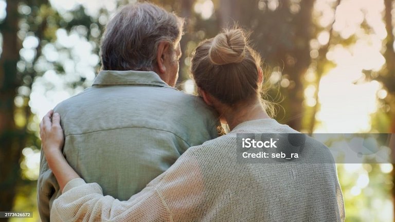 Eine ältere und eine jüngere Person stehen eng umarmt im Wald, von hinten gesehen.