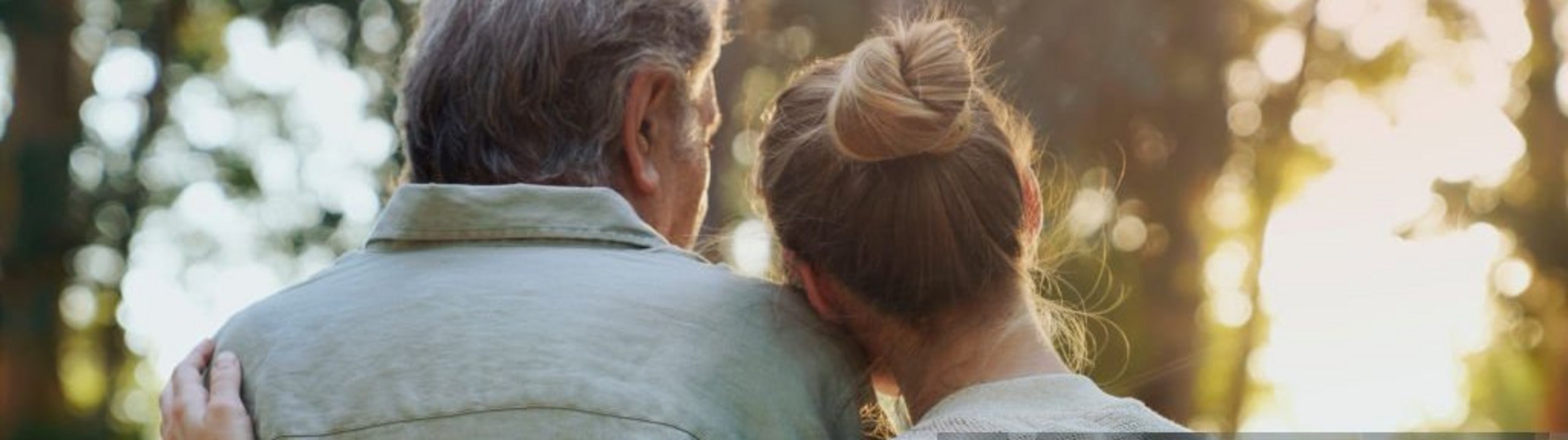 Eine ätere und iene jüngere Person stehen eng umarmt im Wald, von hinten gesehen.