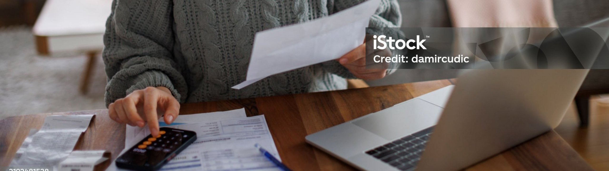 Junge Frau sitzt am Esstisch im Wohnzimmer und sichtet Unterlagen mit Taschenrechner und Laptop.