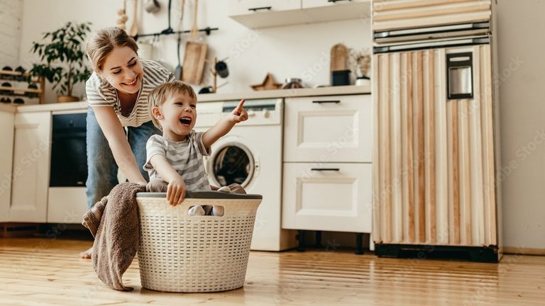Mutter schiebt lachendes Kind im Wäschekorb durch die Küche.