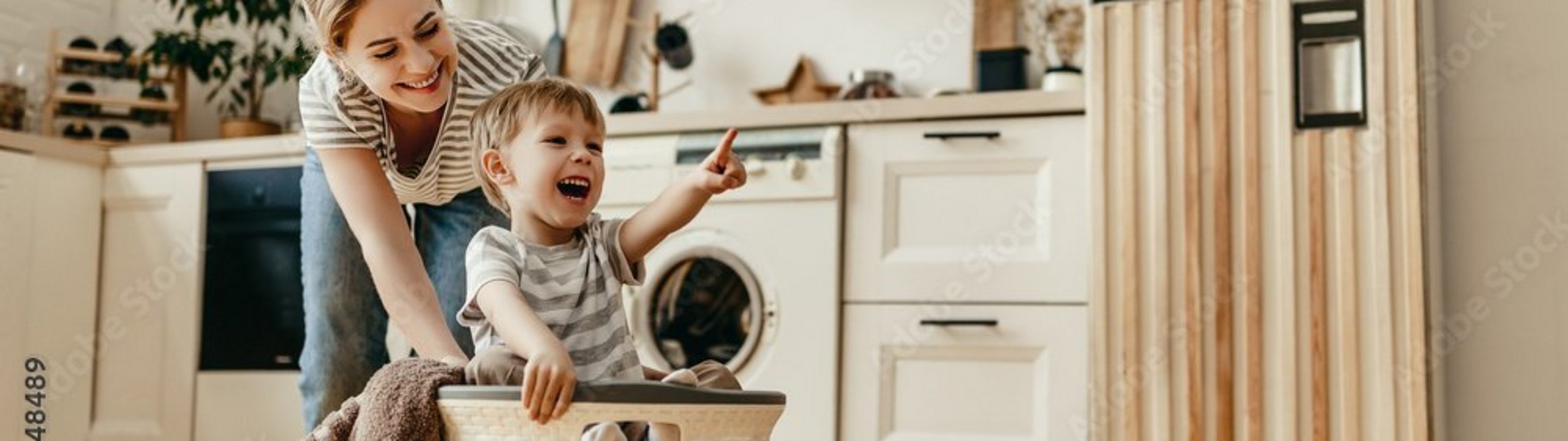 Mutter schiebt lachendes Kind im Wäschekorb durch die Küche.