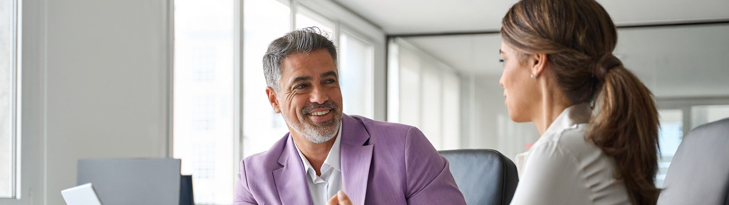 Ein Finanzberater sitzt in einem hellen Büro einer Frau gegenüber, beide sprechen miteinander und sitzen vor einem geöffneten Laptop an einem Schreibtisch.