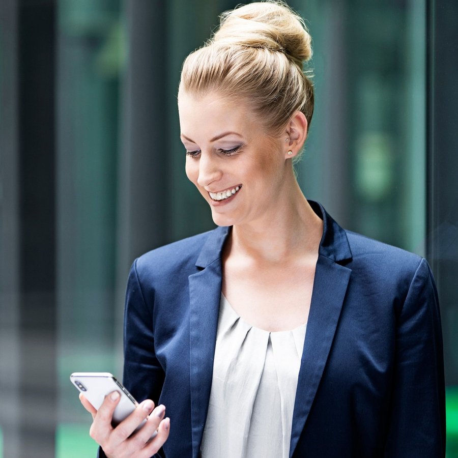 Frau in blauem Blazer schaut lächelnd auf ihr Smartphone.