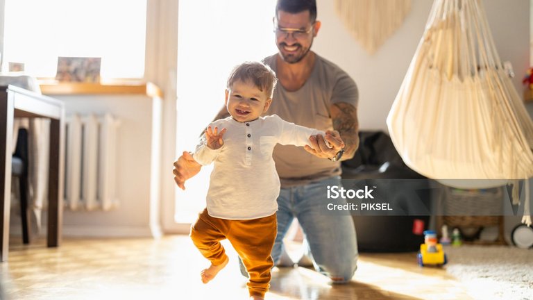 Vater unterstützt lachendes Kleinkind beim Laufenlernen im Wohnzimmer.