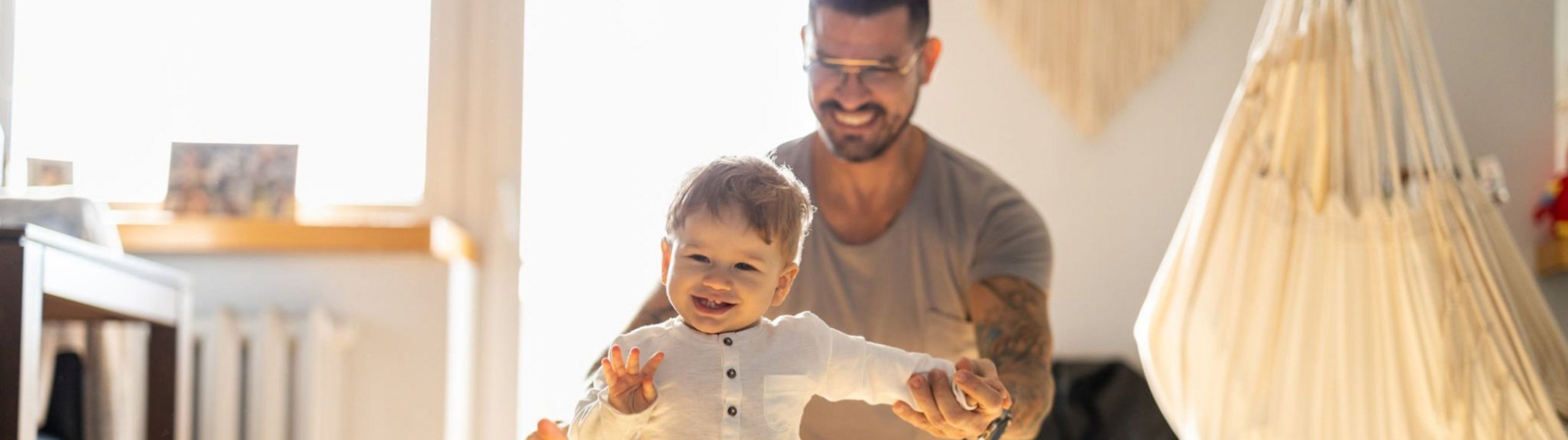 Vater unterstützt lachendes Kleinkind beim Laufenlernen im Wohnzimmer.