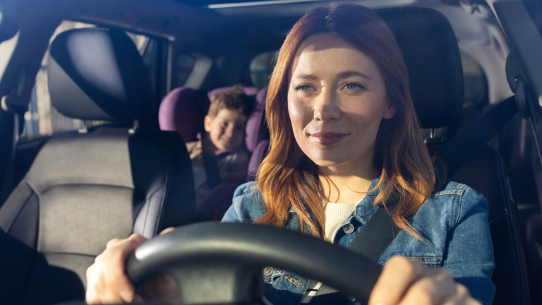 Eine Frau fährt Auto, während ein Kind auf dem Rücksitz sitzt. Beide sind angeschnallt.