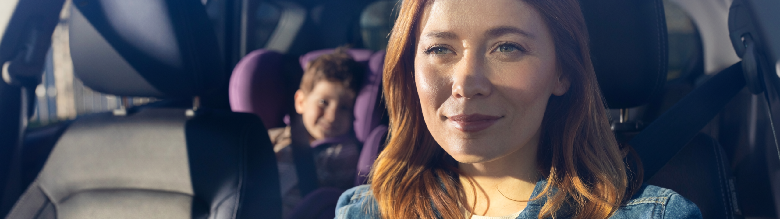 Eine Frau fährt Auto, während ein Kind auf dem Rücksitz sitzt. Beide sind angeschnallt.