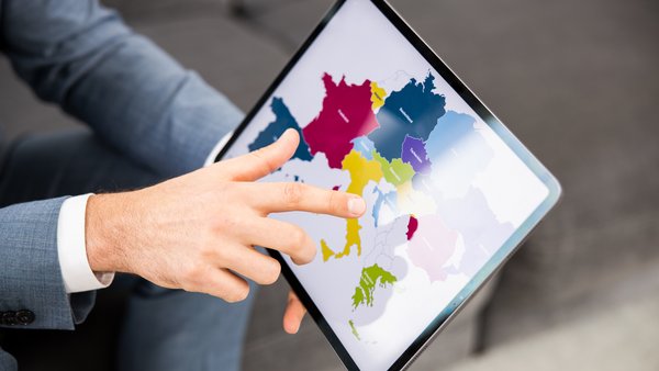 Person hält ein Tablet in den Händen, auf dem eine Europakarte abgebildet ist und zeigt mit einem Finger auf die Europakarte.