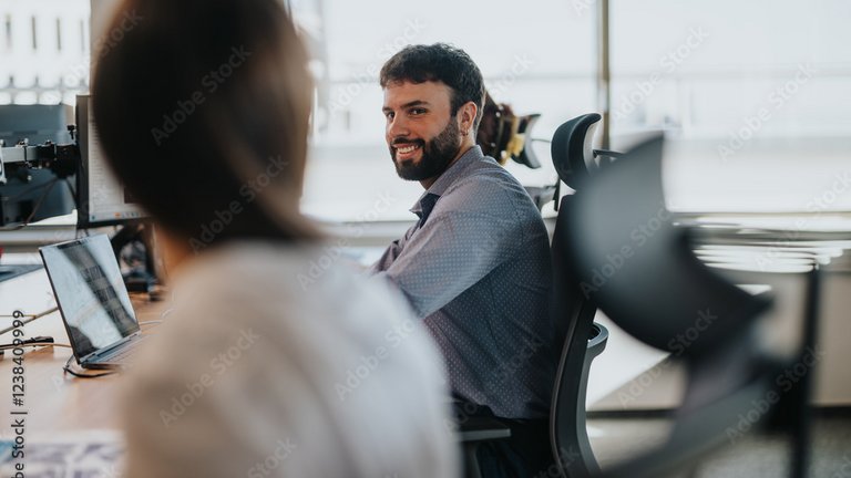 Eine Person arbeitet an einem Schreibtisch im Büro und lächelt einer Kollegin oder einem Kollegen zu.