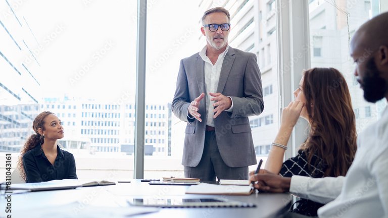 Eine Person präsentiert in einem Besprechungsraum vor einem Team, während die Teilnehmenden aufmerksam zuhören und Notizen machen