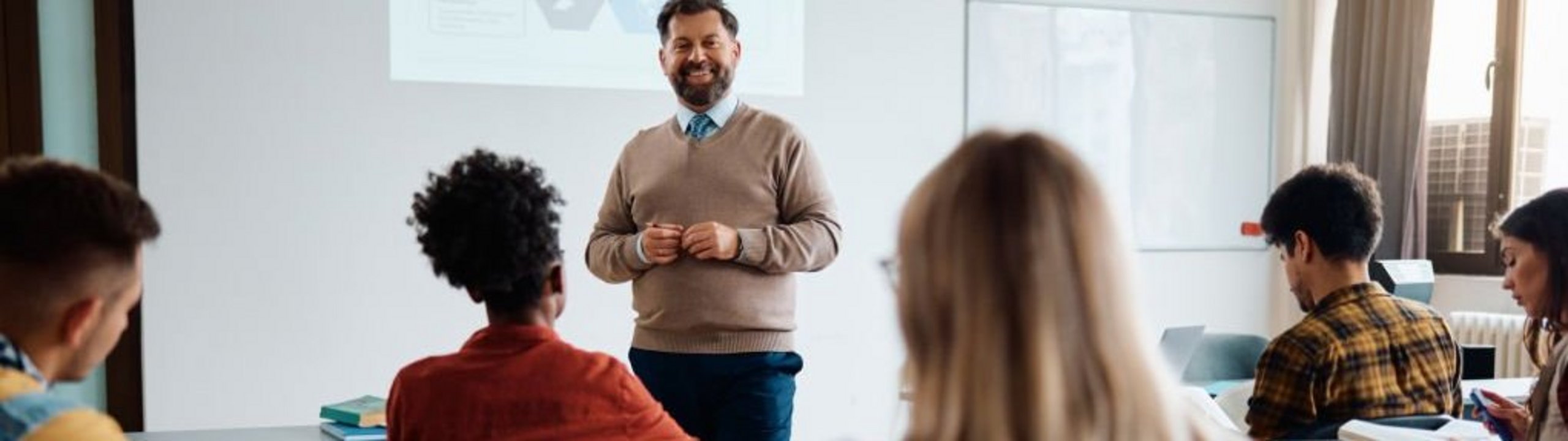 Ein Dozent hält einen Vortrag vor Studierenden in einem Seminarraum, während diese aufmerksam zuhören und Notizen machen.