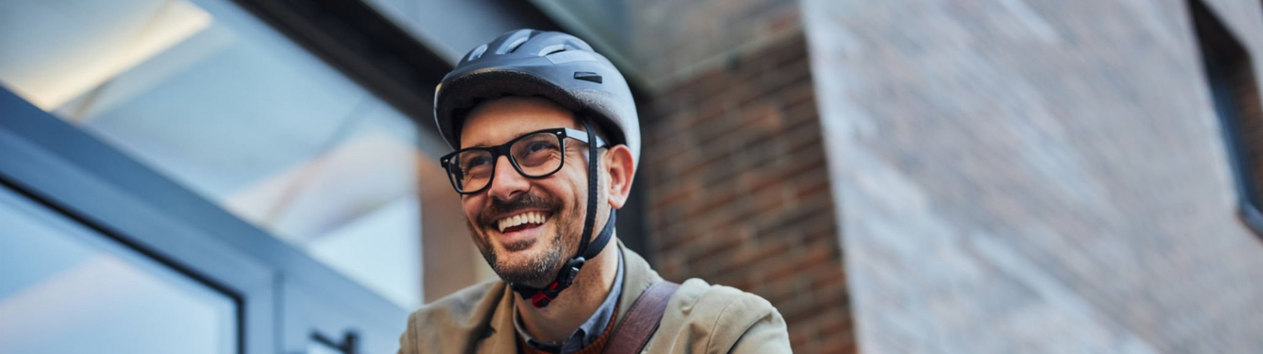 Ein lächelnder Mann mit Brille, Fahrradhelm und beigefarbener Jacke sitzt auf einem Fahrrad vor einem modernen Gebäude.