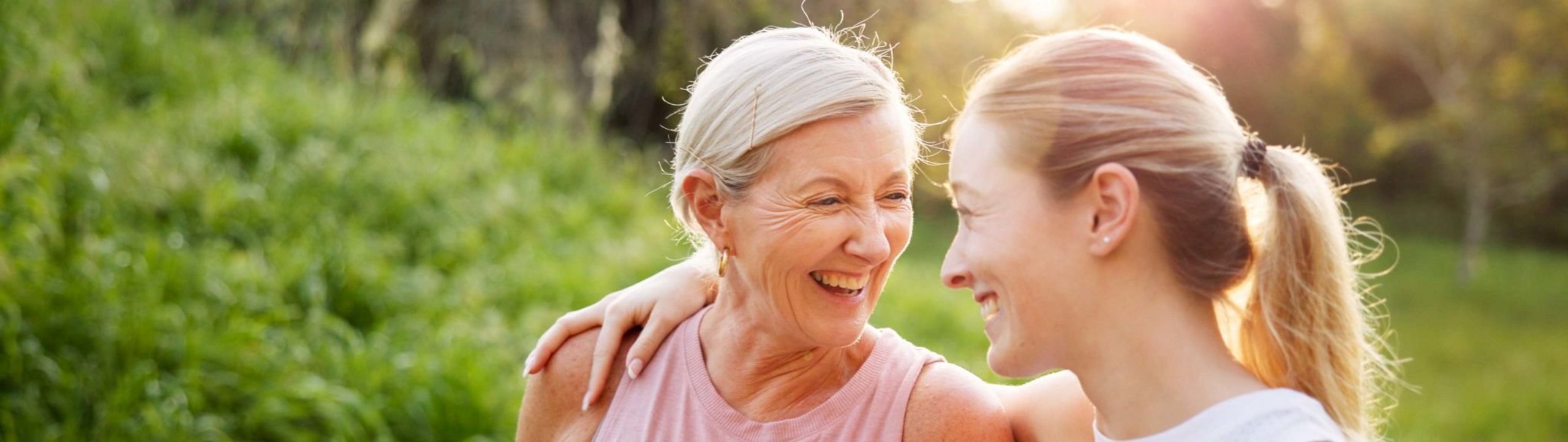 Zwei Frauen unterschiedlichen Alters stehen bei sonnigem Wetter in der Natur, lachen gemeinsam und legen vertraut die Arme umeinander.