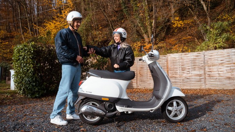 Zwei Personen mit Helmen stehen lachend neben einem Motorroller.