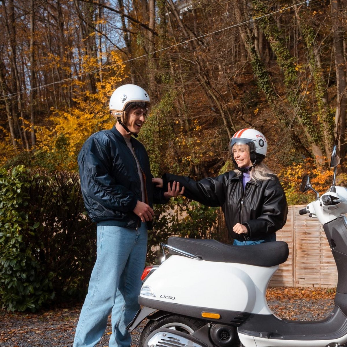 Zwei Personen mit Helmen stehen lachend neben einem Motorroller.