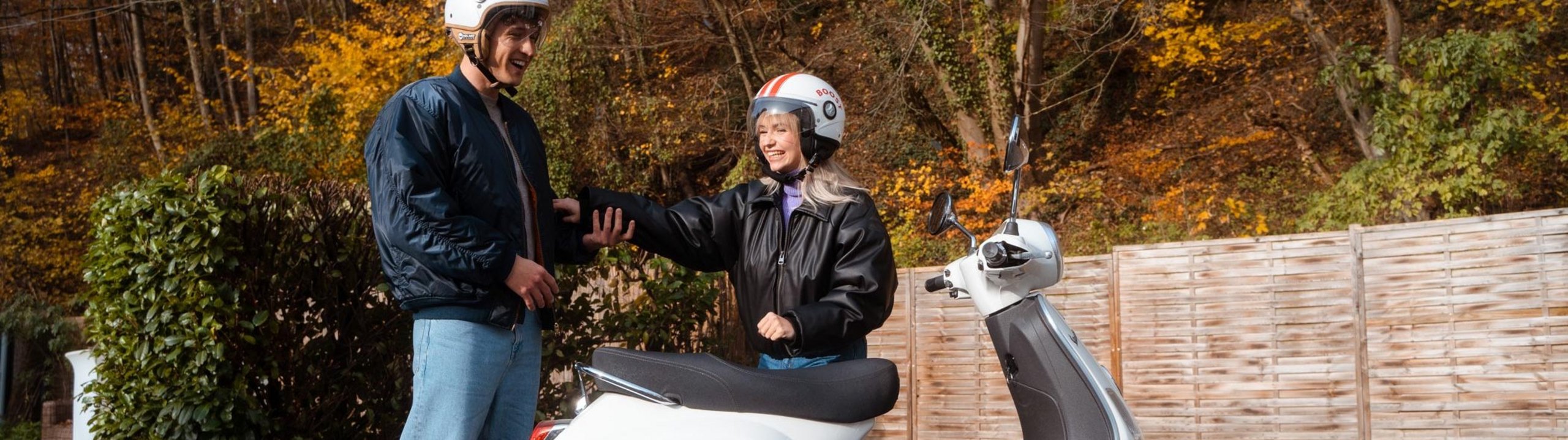 Zwei Personen mit Helmen stehen lachend neben einem Motorroller.