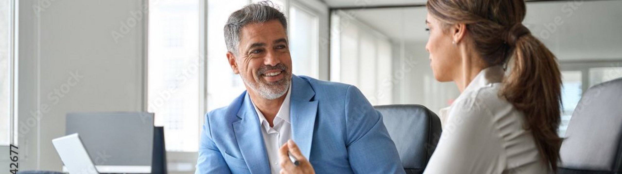 Ein Finanzberater im blauen Sakko sitzt in einem hellen Büro einer Frau gegenüber, beide sprechen miteinander und sitzen vor einem geöffneten Laptop an einem Schreibtisch.