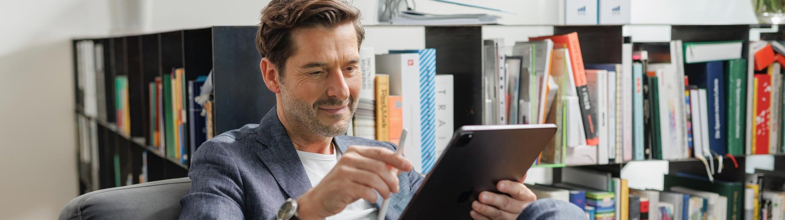 Mann sitzt im Sessel und arbeitet mit einem Tablet vor einem Bücherregal.