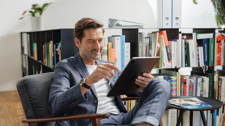 Mann sitzt im Sessel und arbeitet mit einem Tablet vor einem Bücherregal.