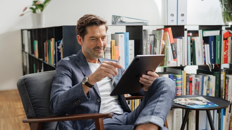 Mann sitzt im Sessel und arbeitet mit einem Tablet vor einem Bücherregal.