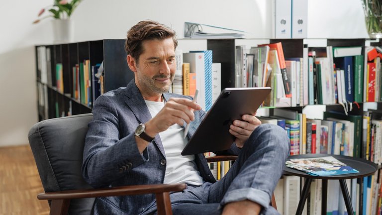 Mann sitzt im Sessel und arbeitet mit einem Tablet vor einem Bücherregal.