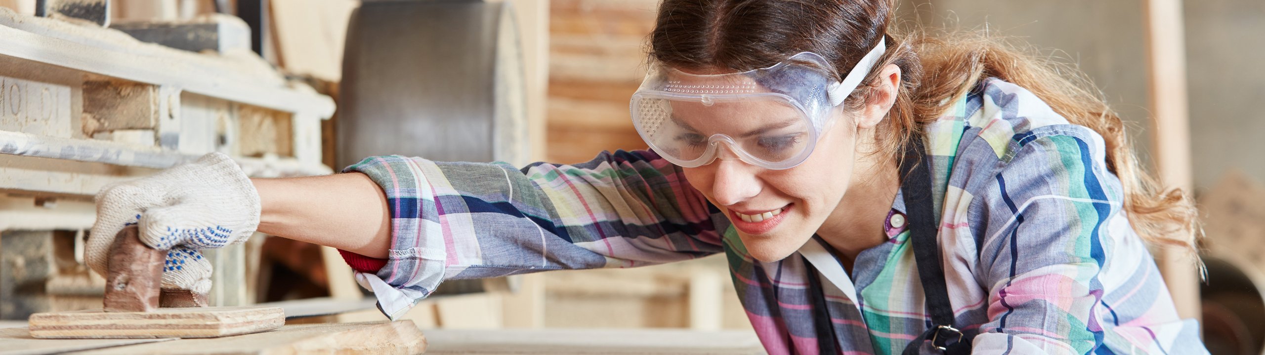 Frau mit Schutzbrille bearbeitet Holz in einer Werkstatt.