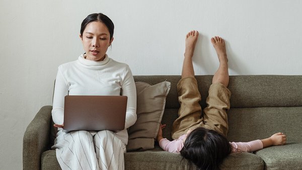 Mother sits on the sofa with a laptop and her daughter lies next to her  