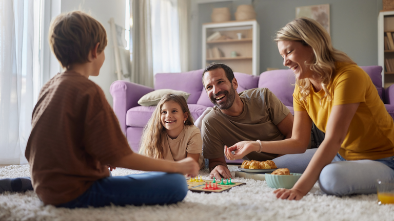 Familie sitzt auf dem Teppich im Wohnzimmer und spielt gemeinsam ein Brettspiel.