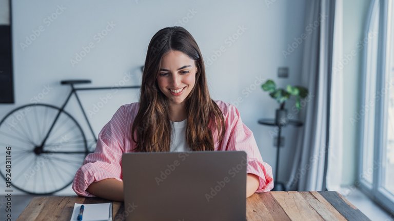 Eine lächelnde Person arbeitet an einem Laptop an einem Holztisch im Homeoffice.