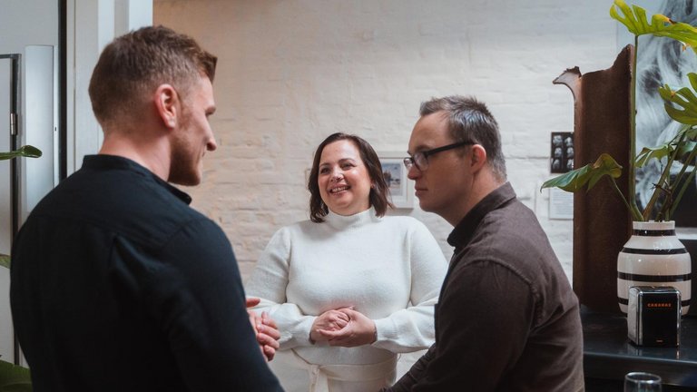 Drei Personen stehen im Gespräch in einem Innenraum und lächeln.