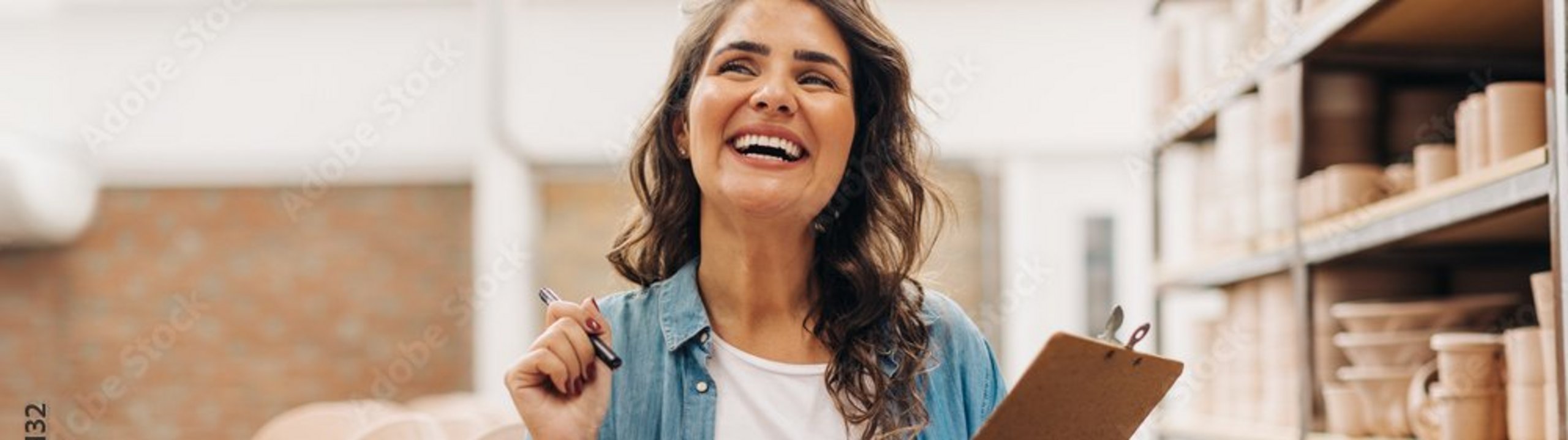 Eine Frau steht lächelnd in einem Geschäft zwischen Regalen mit Keramik und hält ein Klemmbrett sowie einen Stift in der Hand.