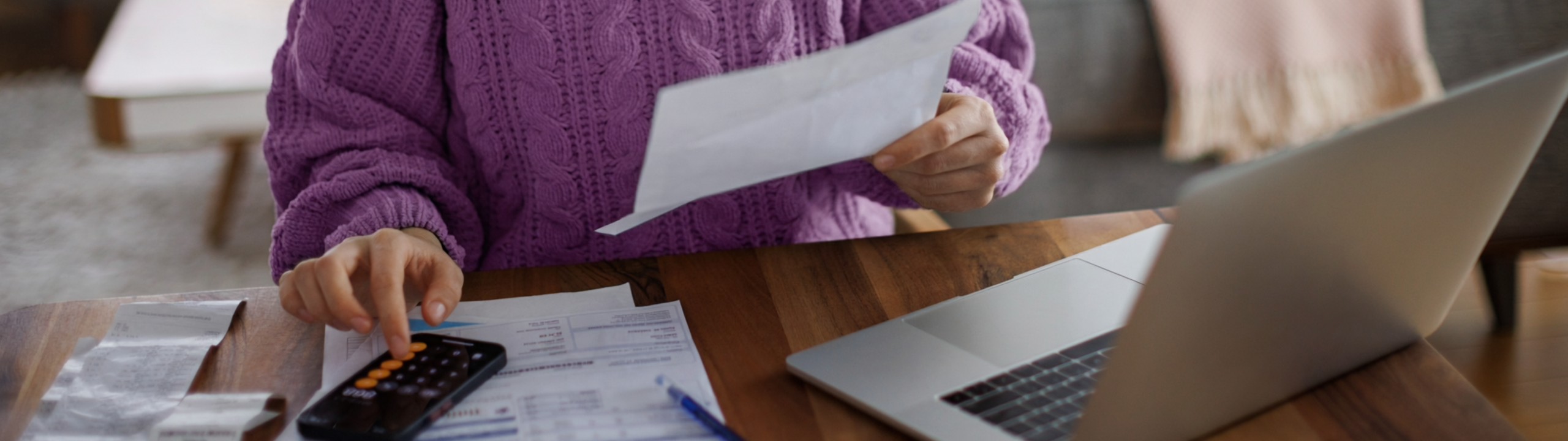 Junge Frau sitzt am Esstisch im Wohnzimmer und sichtet Unterlagen mit Taschenrechner und Laptop.