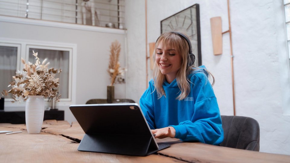 Junge Frau mit Kopfhörern arbeitet lächelnd an einem Tablet am Tisch.