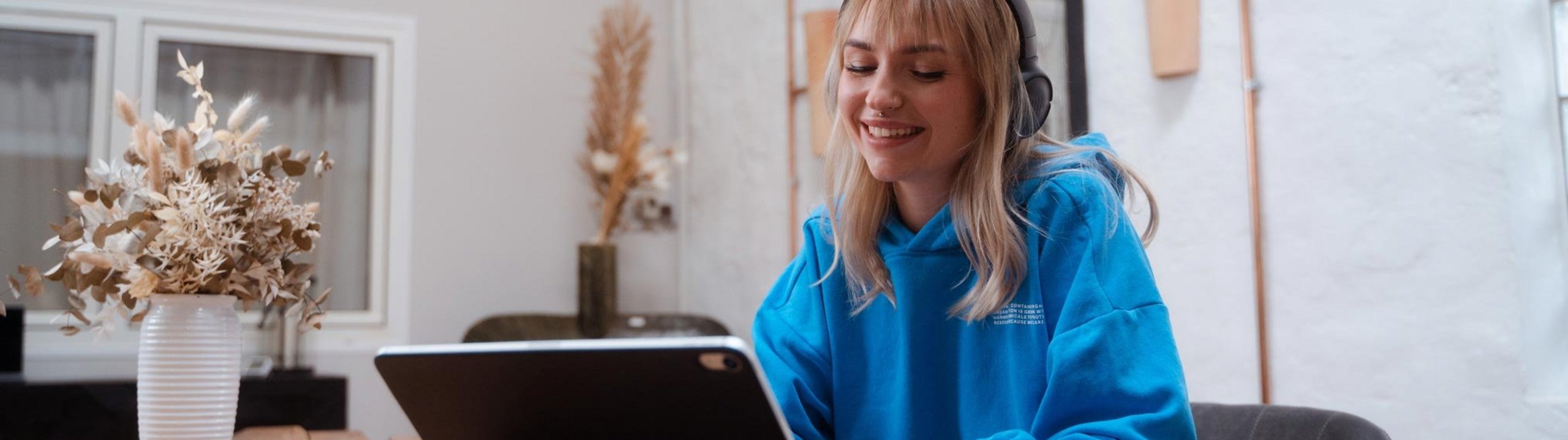 Junge Frau mit Kopfhörern arbeitet lächelnd an einem Tablet am Tisch.