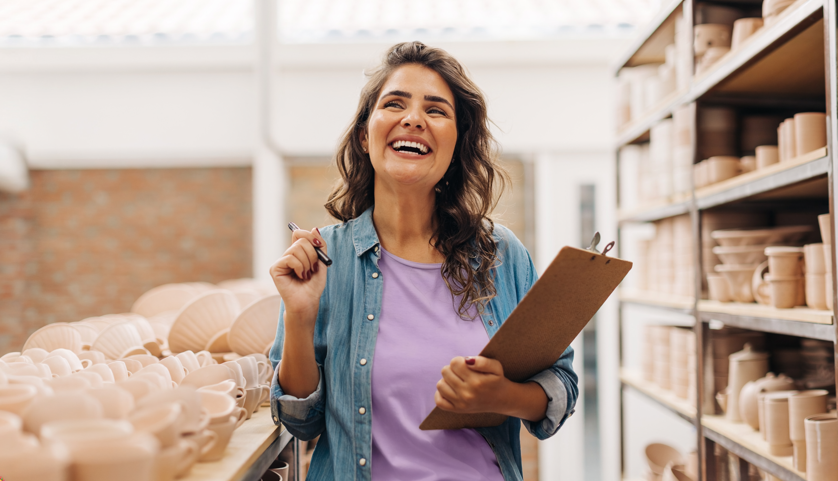 Eine Frau steht lächelnd in einem Geschäft zwischen Regalen mit Keramik und hält ein Klemmbrett sowie einen Stift in der Hand.
