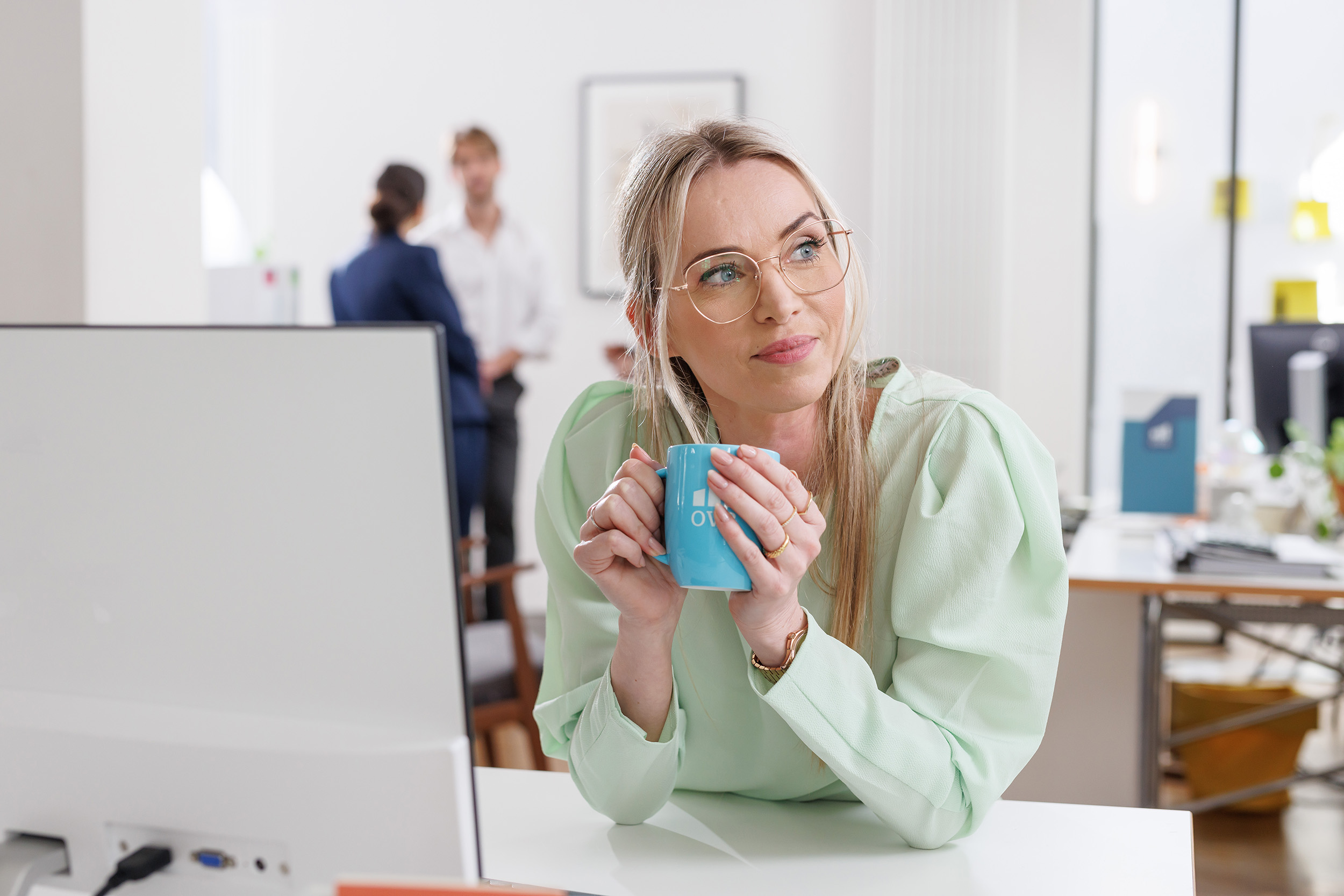 Eine Frau sitzt vor ihrem Arbeitsplatz mit einer Tasse Kaffee und schaut zuversichtlich