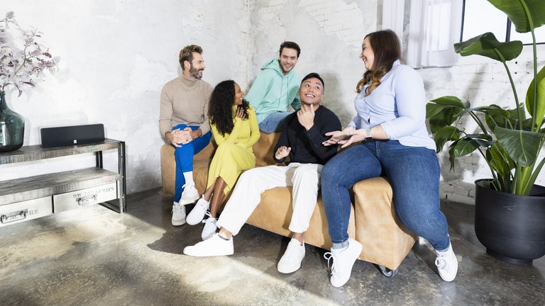 Eine Gruppe von fünf Personen sitzt gemeinsam auf einem Sofa und unterhält sich lachend.