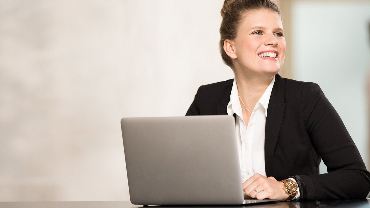 Frau in Business Kleidung sitzt vor einem Laptop und lächelt.