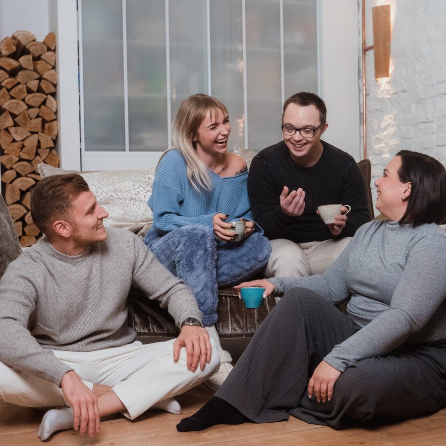 Four young adults are sitting in a cosy living room, laughing and chatting with cups of coffee in their hands.