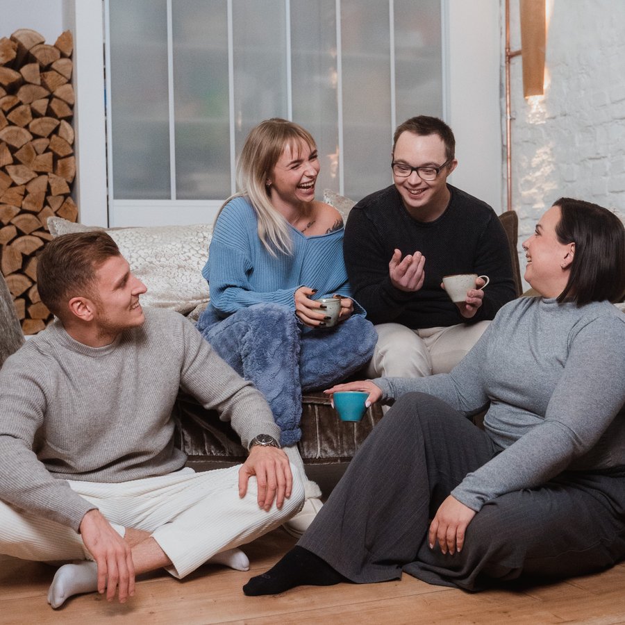 Vier junge Erwachsene sitzen lachend mit Kaffeetassen in einem gemütlichen Wohnzimmer und unterhalten sich.
