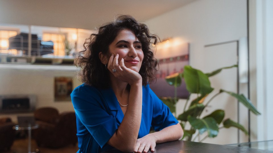Junge Frau in blauer Bluse sitzt an einem Tisch und blickt nachdenklich grinsend in die Ferne.