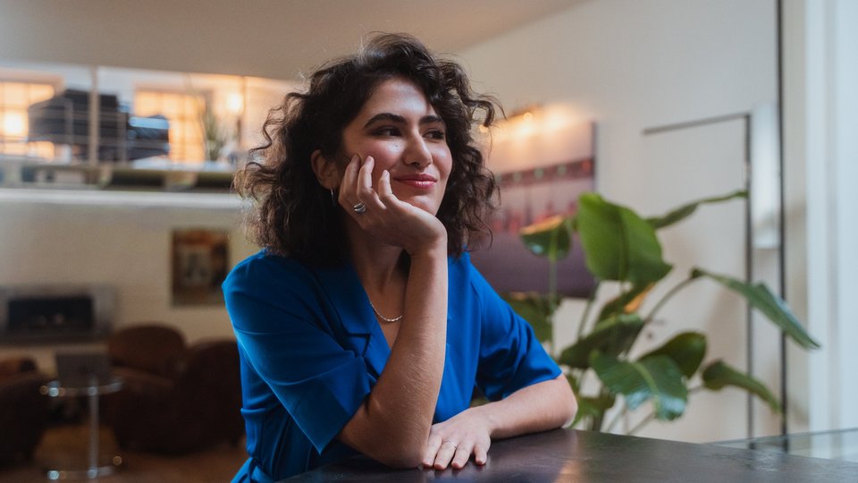 Young woman in a blue blouse sits at a table, gazing into the distance with a thoughtful smile.